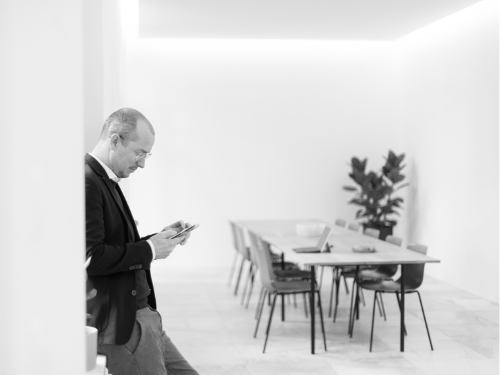 Ein Mann lehnt an eine Wand mit Blick auf das Smartphone in einem hellen Meetingraum.