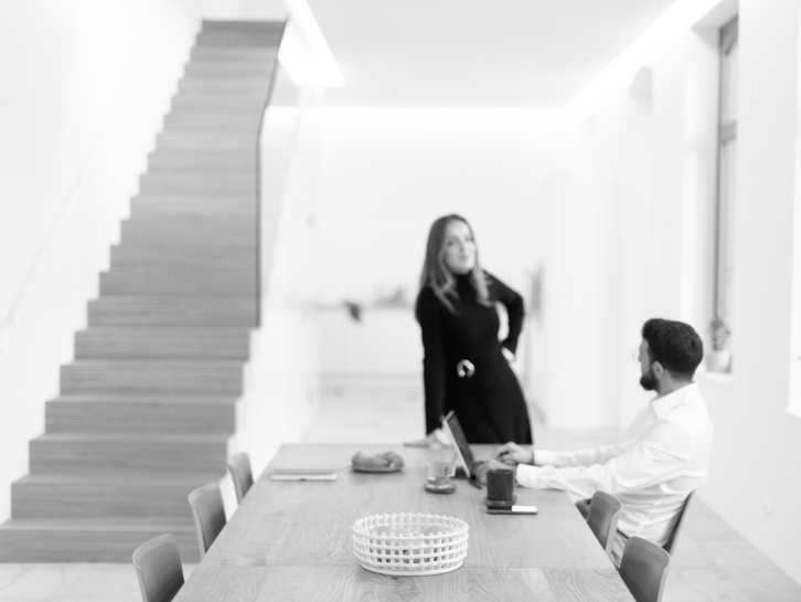 Two people in a modern, bright office; a man sits at a table with a laptop, a woman stands next to him, in the background near the stairs.